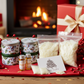Christmas-themed products including candles, oils, and packaging on a table with a fireplace in the background.